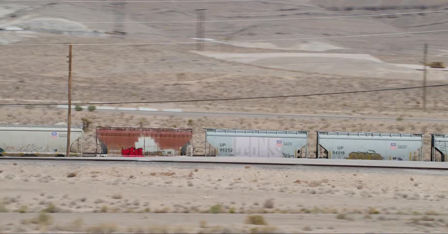 A landscape of bare mountains in the background and a motionless freight train alongside a highway with traffic in between but the focus is on the freight train we can also see telephone poles wires and another road