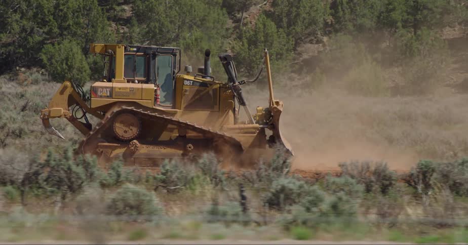 A tractor drives in some dirt in front of several trees as cars pass by
