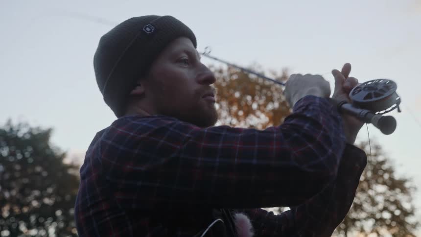 Man with black beanie beard and plaid shirt casts a fishing rod