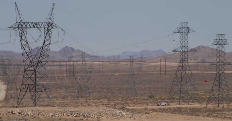 A field of many interconnected pylons or utility poles holding power lines towering over a vehicle on a dirt road with mountains off in the distance