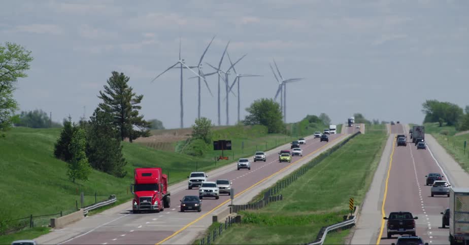 Traffic is shown driving on the highway past some grassland with wind turbines