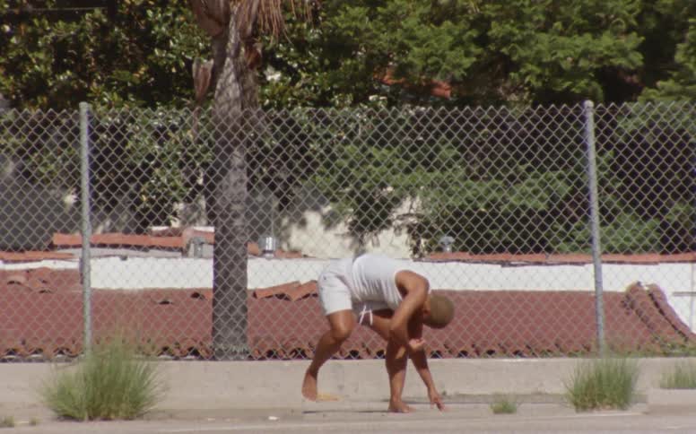 Woman with short hair white tank top and shorts dances in front of mesh fence at the park