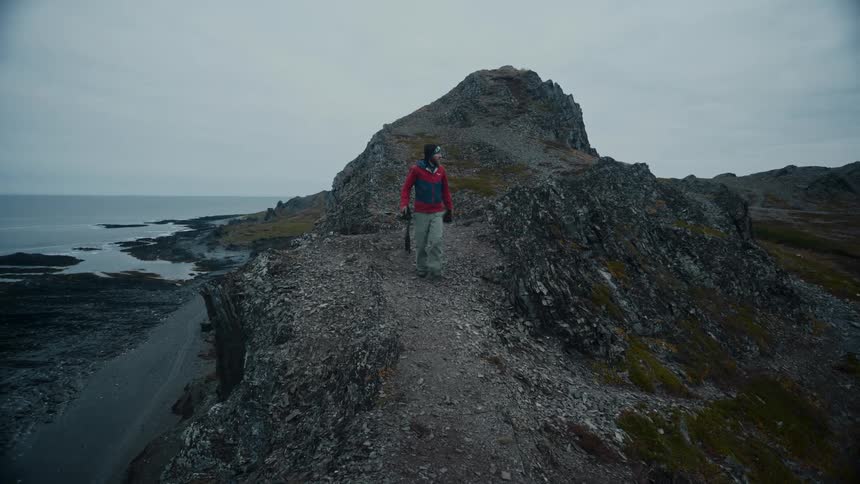 Man wearing red coat and gray pants walks downhill carrying a camera on the shore