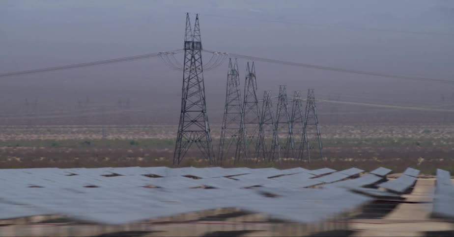 There is a row of 7 utility poles with power lines connecting them with mountains in the background on the horizon and small roofed structures in the foreground the land is flat and brown