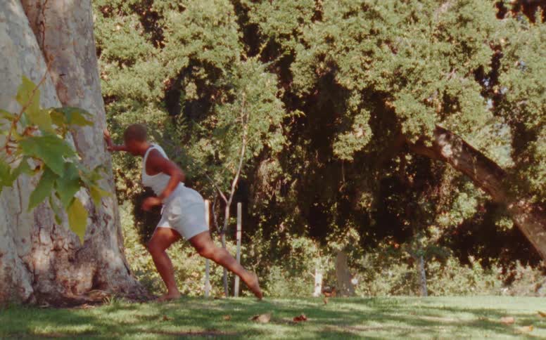 Female dancer with short hair white tank top and shorts dances near thick tree trunk at the park
