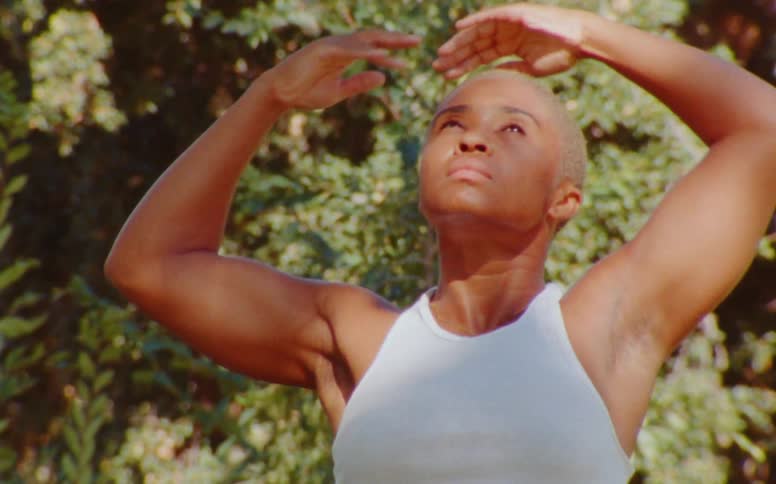 A brownskinned woman with shaved head performs a sequence of intricate arm hand motions while gazing serenely at the sky or with downcast eyes in front of some bushes