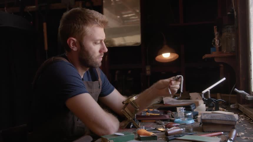 Clockmaker with beard and dark blue t shirt holds wristwatch sitting at table