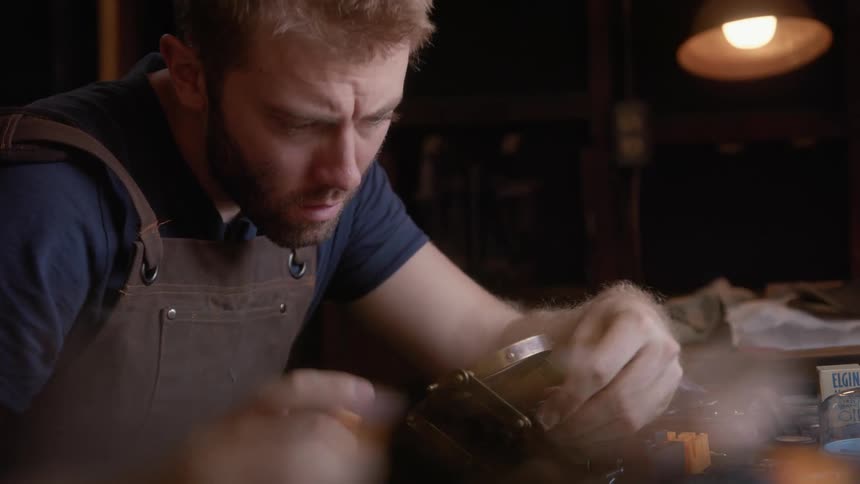 A man wearing an apron repairs a piece of jewelry on a table full of tools