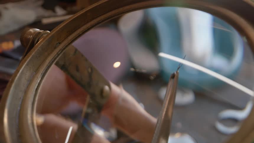 A watchmaker uses tweezers to inspect a small part under a magnifying glass