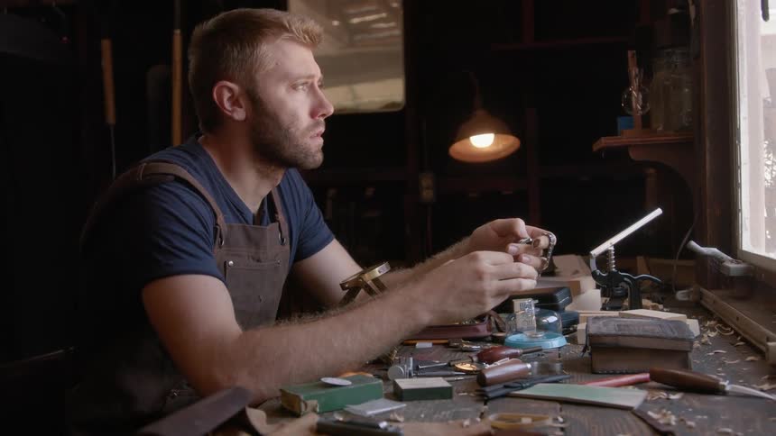 Clockmaker with beard works at desk thinking