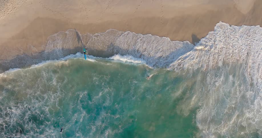 Clear water waves roll into shore and over the beach as a surfer carries their surfboard into the water and rides