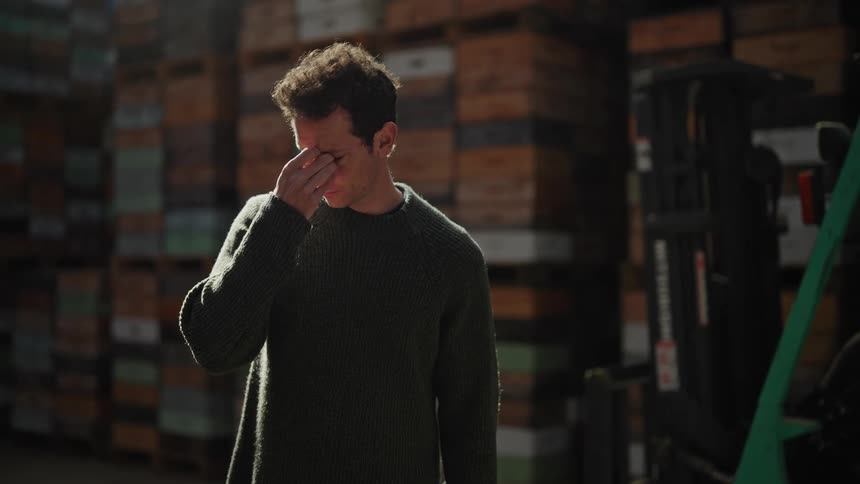 A man is standing in a room with stacks of boxes as he rubs his eyes with his hand and fingers