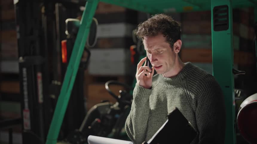 A man is standing holding a notebook and talking on the phone as he shakes his head