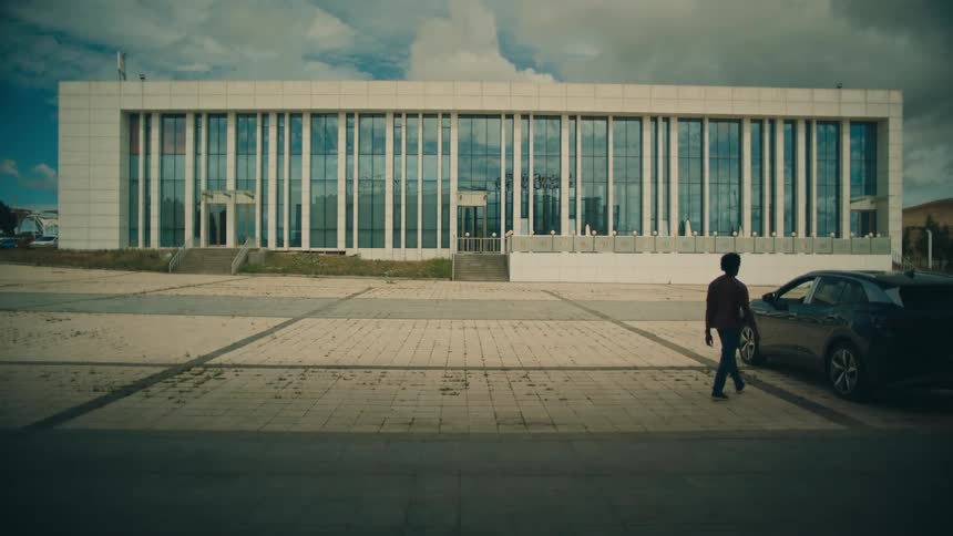 Building reflects the sky as man walks across parking lot and opens car door