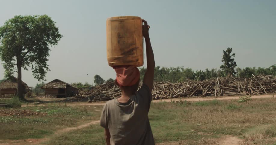 Woman wearing red hat and dirt t shirt carries bucket on her head in the village