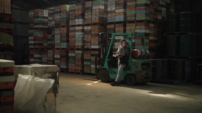 Man in gray sweater leans against the forklift talking on smartphone at the warehouse