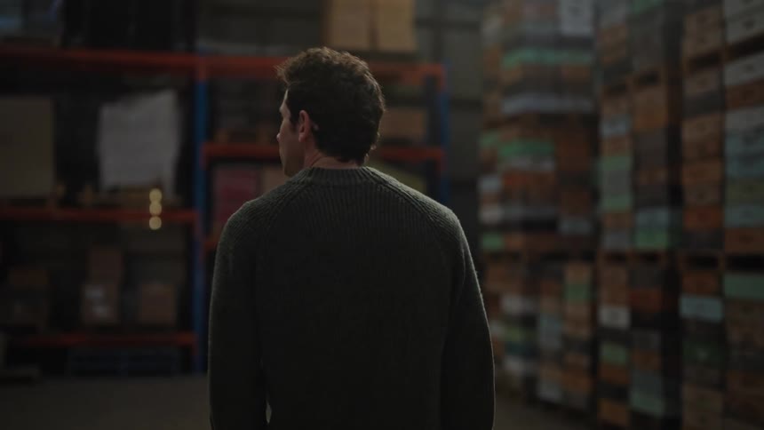 A man walks through a dimly lit warehouse full of shelves stacked from floor to ceiling with boxes he looks around as he walks