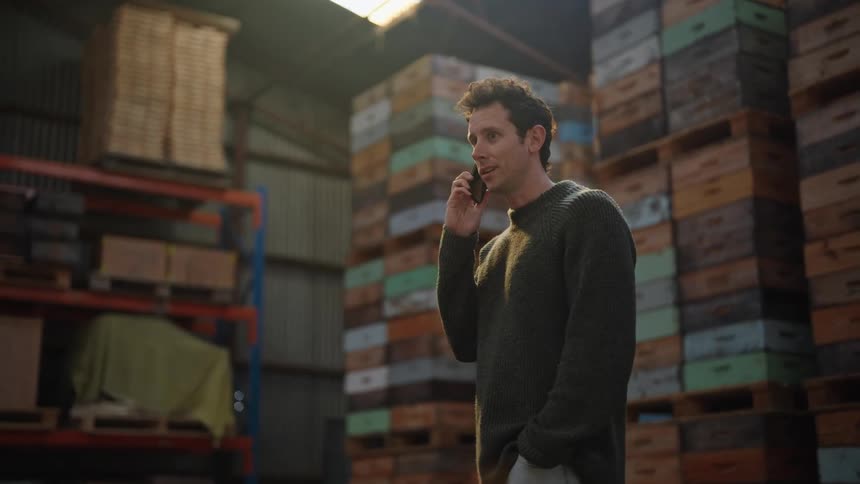 Man talks on smartphone while standing in warehouse aisle near large stacks of merchandise