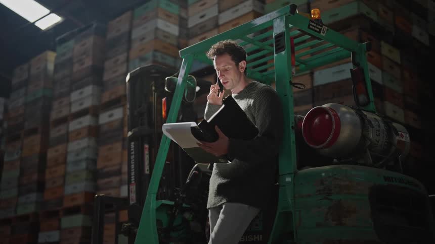 Man in gray sweater leans against the forklift talking on smartphone at the warehouse