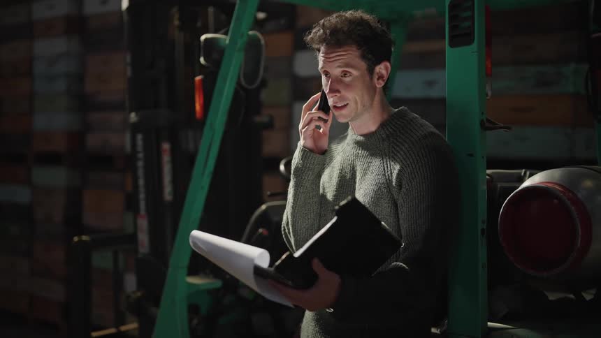 A man holds an opened binder with a clipboard in his left hand talks nodding on the phone held in his right hand while standing in a warehouse in front of loaded shelves stacked with boxes