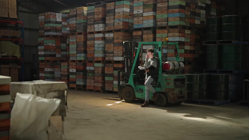 Man in gray sweater leans against the forklift talking on smartphone at the warehouse