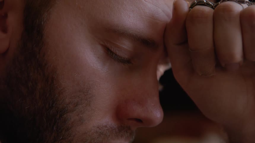 A man with a beard closes his eyes and brings his hand to his forehead in thoughtful tones