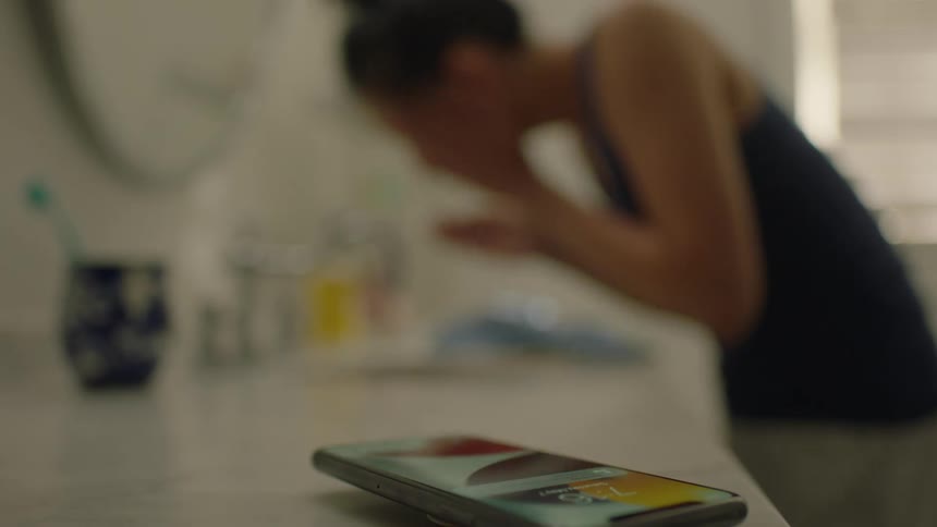 A woman is washing her face in the bathroom while her cell phone rings