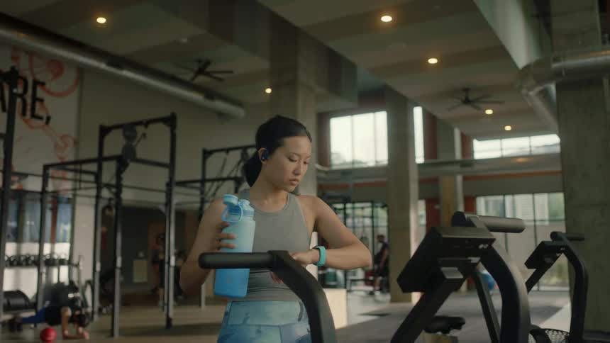 A woman is drinking water from a large blue bottle at the gym