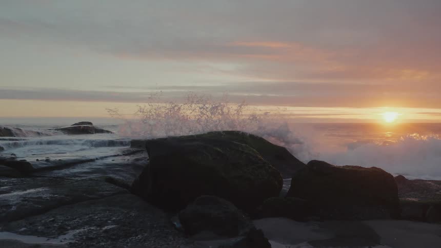 A view of waves crashing against black rocks with the sunrise in the background