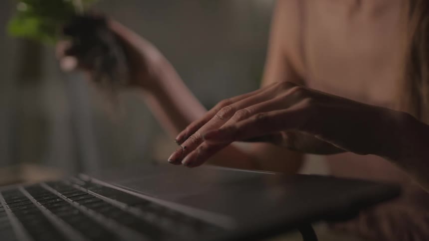 A woman holding a bunch of green leaves with roots dirt attached in one hand scrolls on her laptop with her other hand perhaps looking something up online