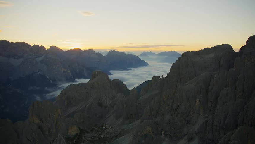 An aerial view of a mountain range and cliffs above the cloud level with the sunrise on the horizon