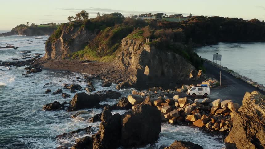 A mini van pulling on the side of the road filled with rocks close to a riverbank as the waves from the river continue to splash on the rocks