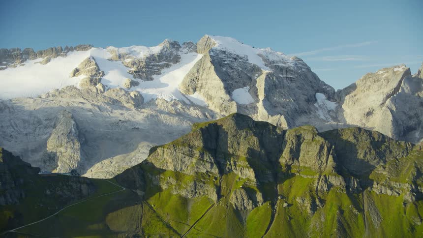 An aerial view of a mountain peak with grass and in the background a larger mountain with its snowcovered peak