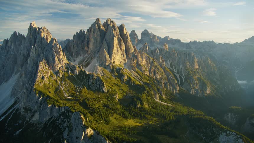 An aerial landscape of a mountain range with geological peaks and coniferous trees around during sunrise