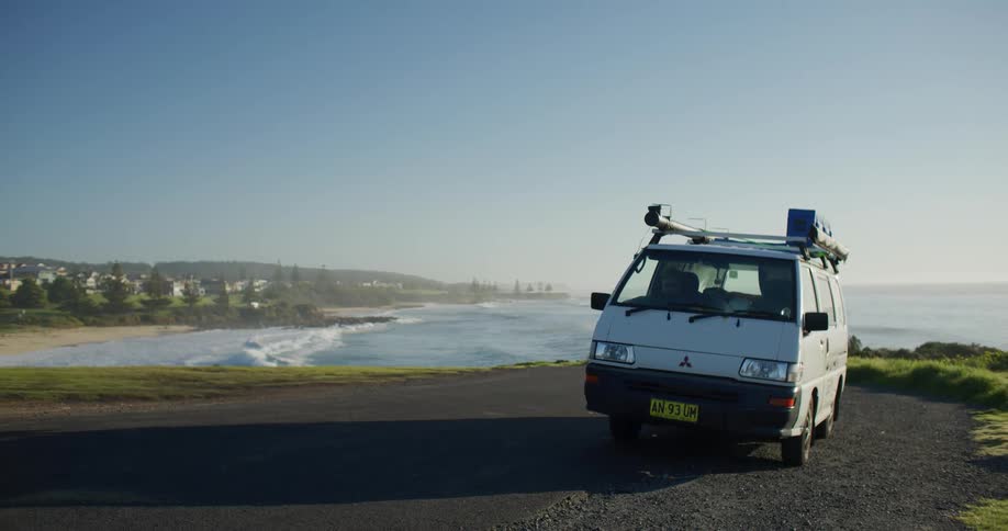 A small van parked and in the background a beach with several waves crashing on the shore