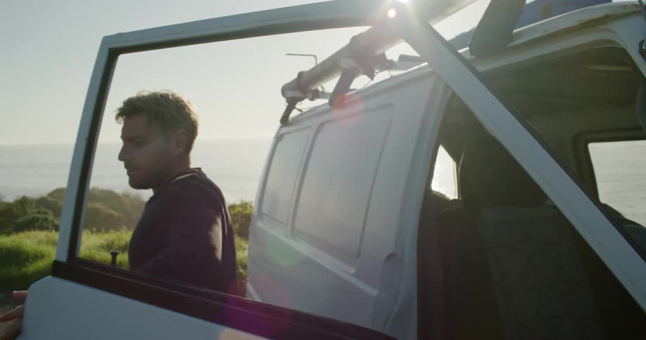 A man gets out of a van and walks behind it and in the background there is a sea