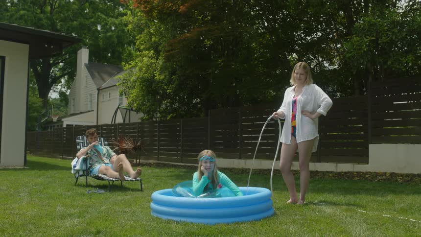 A woman is filling a small pool using a hose with a little girl waiting and a man sitting at the back