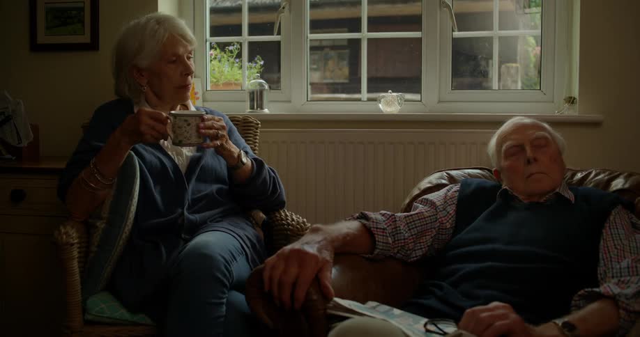 An elderly woman holding a mug looks out the window before touching her sleeping husbands hand