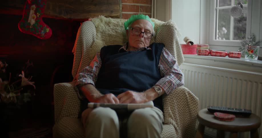 An elderly man wearing a green paper Christmas hat dozes in an armchair