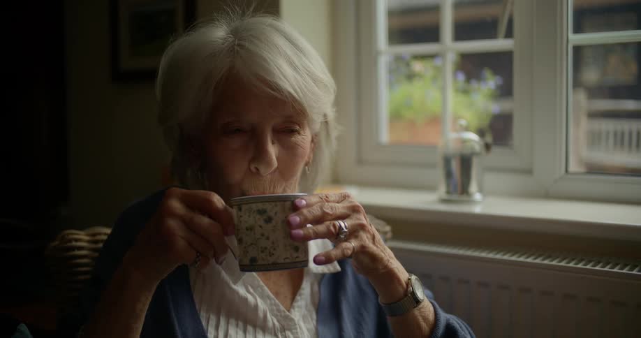 Elderly woman sits on a chair indoors drinking from a mug