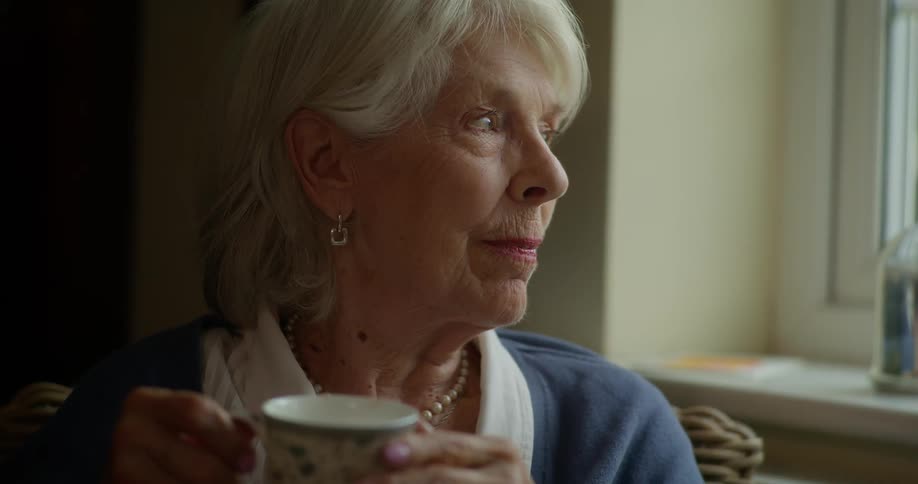 Elderly woman sits next to a window with a cup