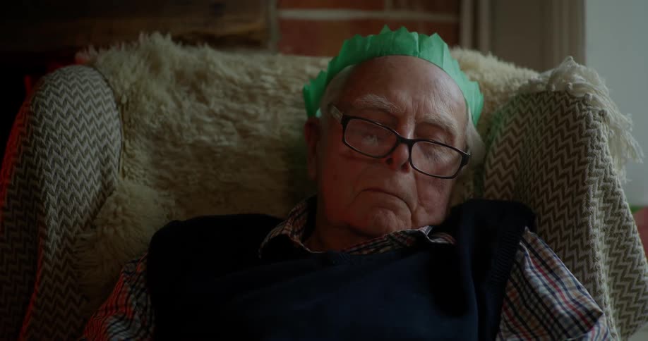 An elderly man with a green paper Christmas hat and glasses dozes in an armchair