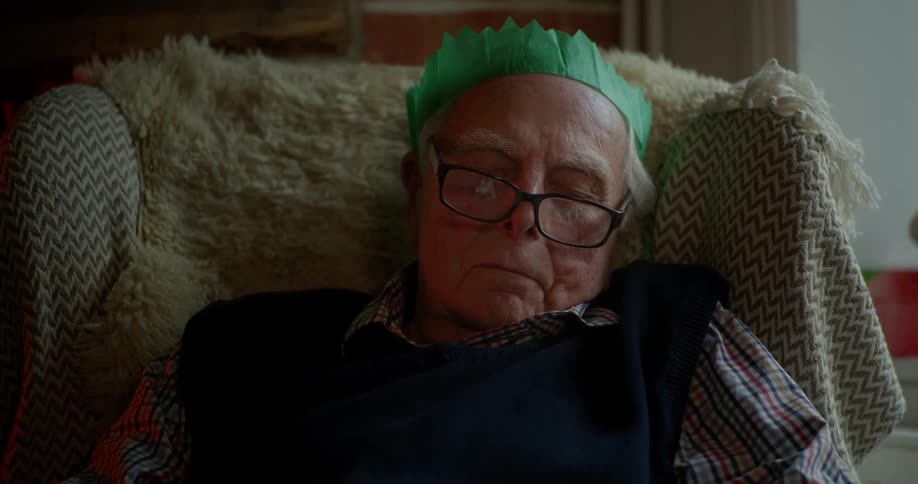 An elderly man wearing a green, paper Christmas hat is sleeping in an armchair