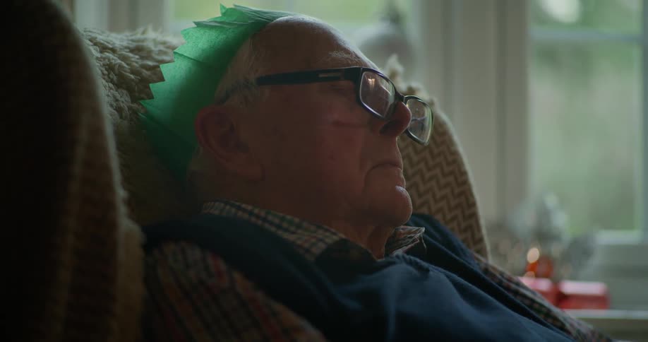 An elderly man wearing a green paper Christmas crown sleeps peacefully in an armchair in a cozy living room