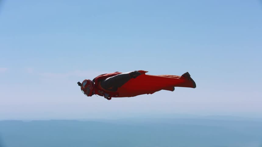 A person in an orange wingsuit floats through the air under a blue sky parallel to the ground