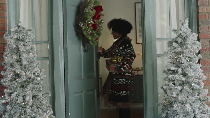 A woman holding a present and entering a boys house who opens the door during christmas time