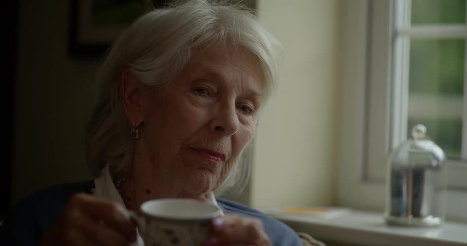 An old woman in partial darkness looks out a window while holding a tea mug