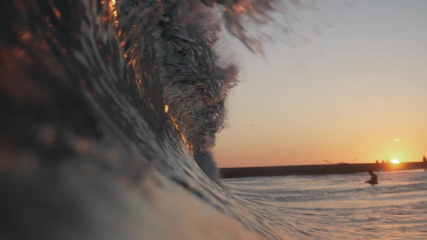 A wave splashes as it curls and rolls into shore and overhead as people on the beach are silhouetted by the setting sun