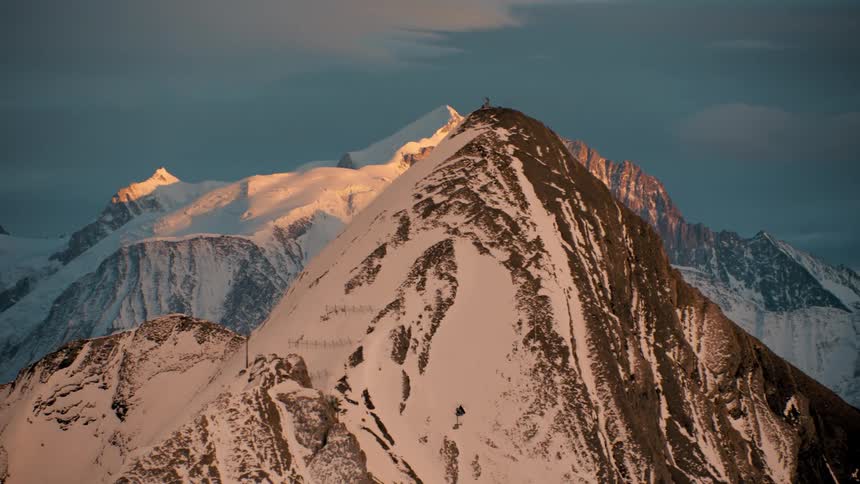 Sunlight shines upon snow covered mountains with a building high on top of a peak