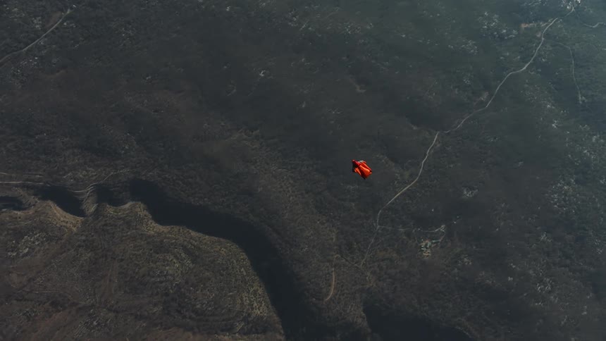 Aerial view above a wingsuit glider moving through sky above a river valley gorge and village during the day
